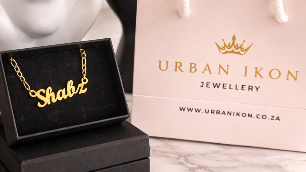 Gold necklace with 'Shabz' on a black box, next to an Urban Ikon Jewellery bag on a marble surface.