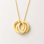 A gold necklace with three interlocking circular rings on a delicate chain, featuring the engraved words 'HONEY', 'BABY', and 'ANGEL' on the rings, set against a clean off-white background.