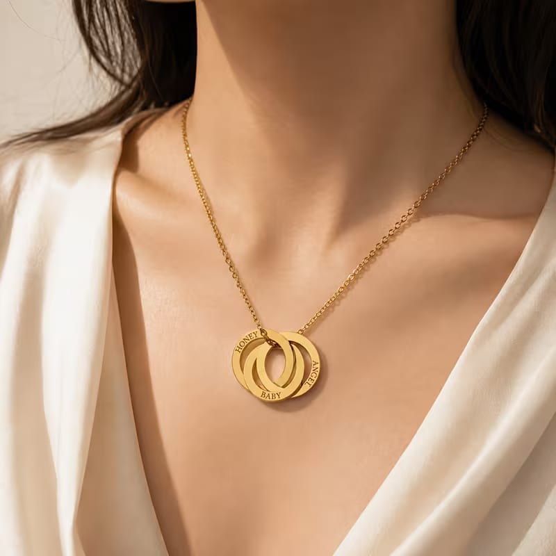 A close-up of a woman wearing a gold necklace with two interlocking circular pendants, one engraved with the name 'ELIZABETH'.